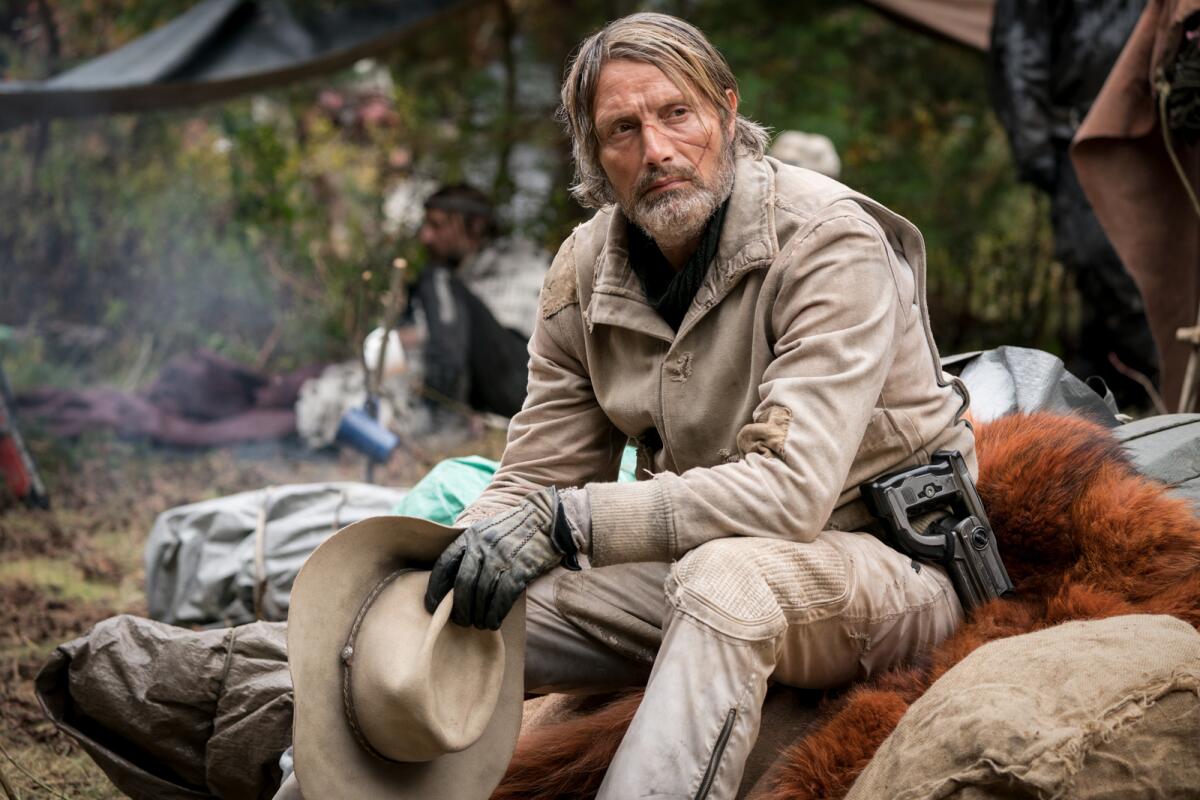 Mads Mikkelsen seated, holding a hat.