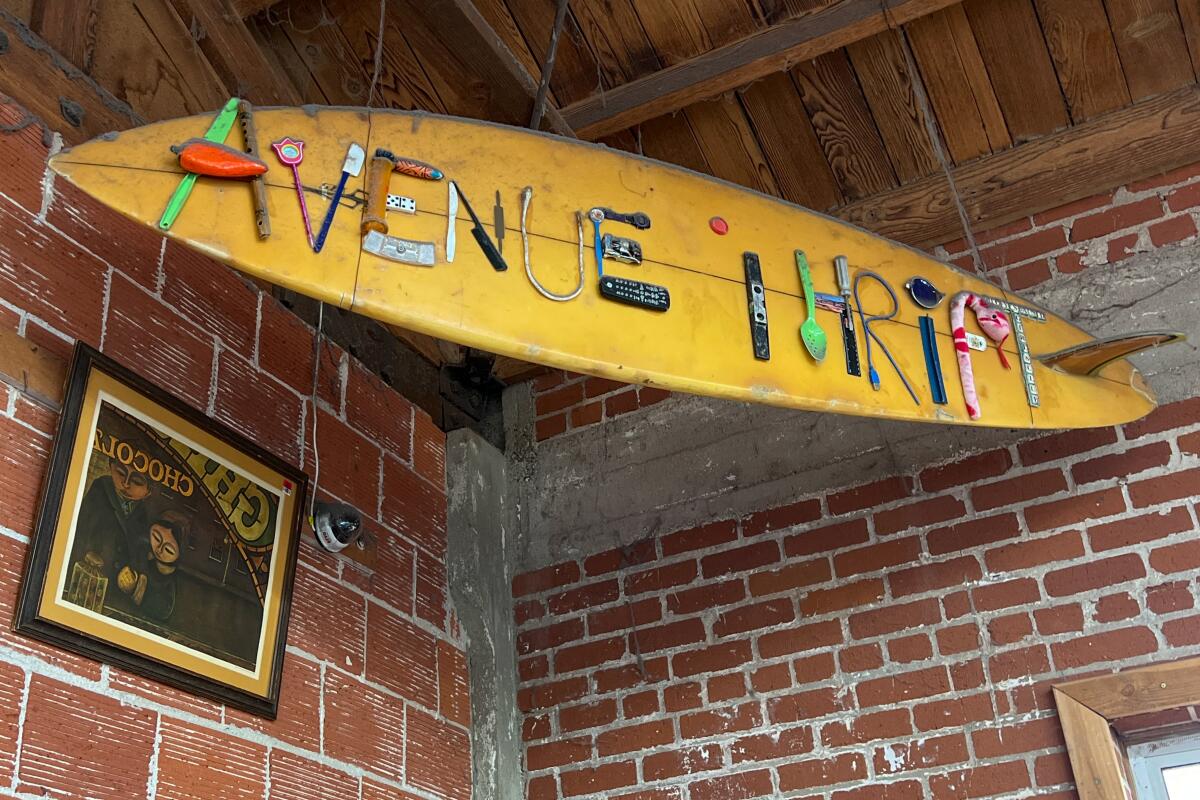 A surfboard with the words Avenue Thrift spelled out in kitchen implements, dice and other items hangs over brick walls.