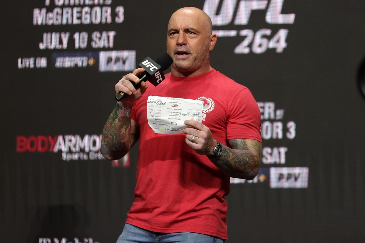 Joe Rogan speaks holding a microphone and a piece of paper while standing on a stage