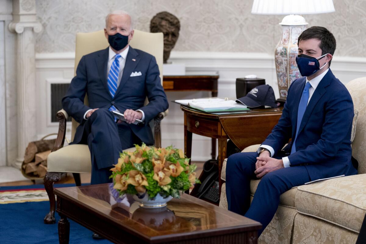 President Biden sits on a chair and Pete Buttigieg sits on a sofa in the Oval Office