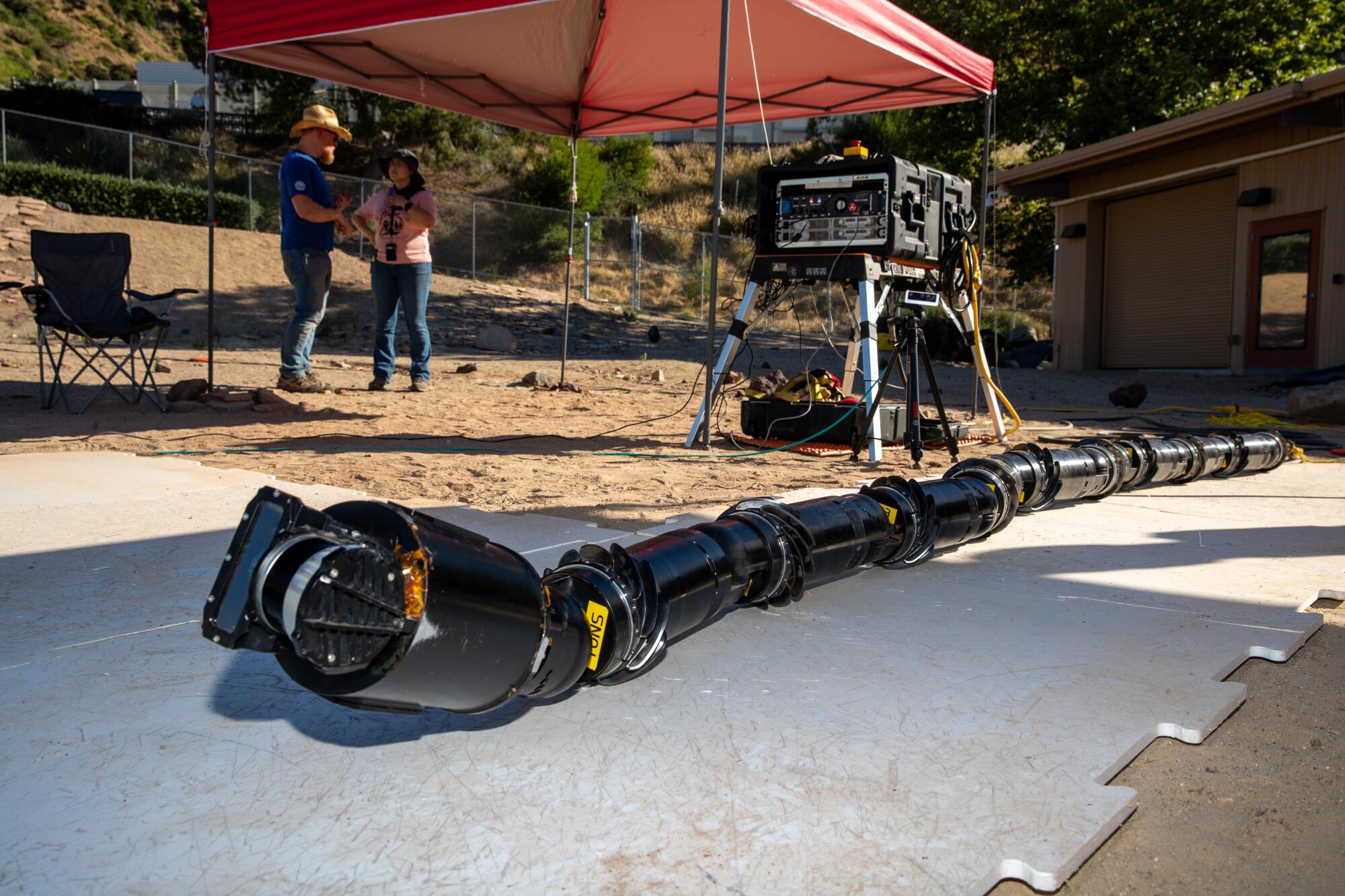 A long, black tube-like object lies on the ground, with two people standing in the background.