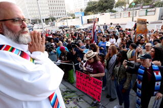 A Catholic priest in white robe looks out over a crowd in downtown Los Angeles.