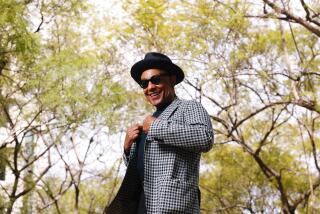 Giancarlo Esposito tugging at the lapels of a black-and-white blazer, wearing a black hat and dark glasses in front of trees