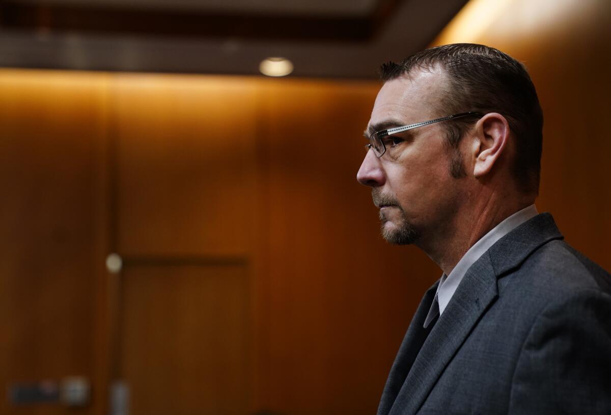 A man in glasses and a suit walks into a wood-paneled courtroom.