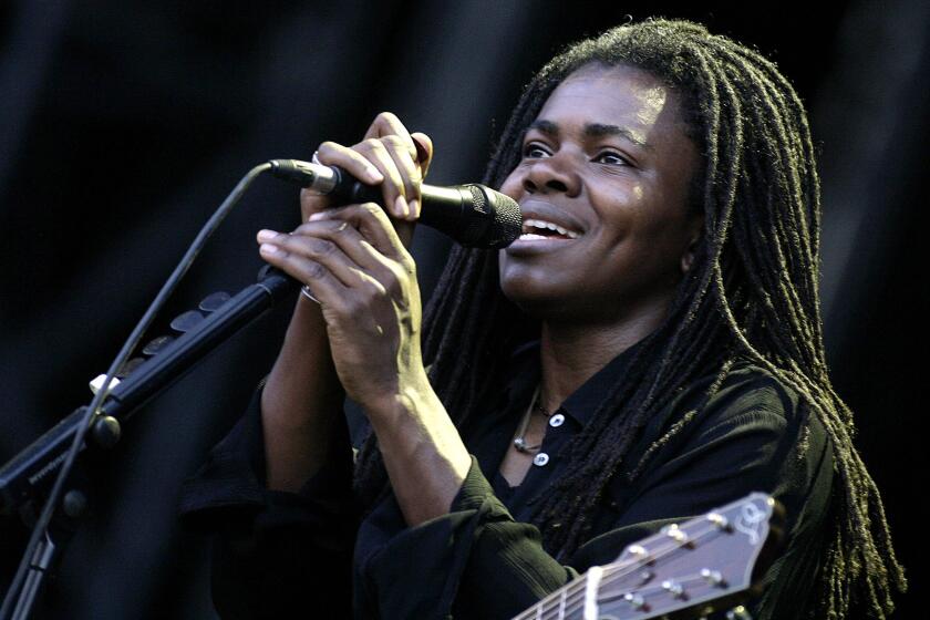 A woman with long dreadlocks wearing a black long sleeve shirt singing into a microphone while a guitar hangs on her body
