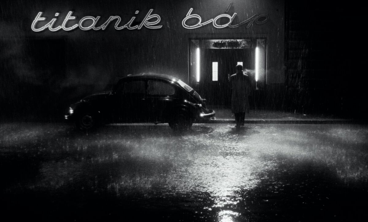 Amid a downpour, a small car sits outside a darkened bar with the neon sign "titanik bar."