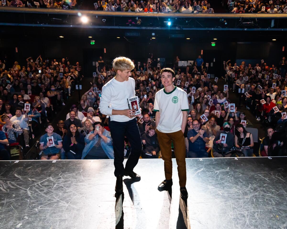 Two people stand on a stage with a full theater behind them.