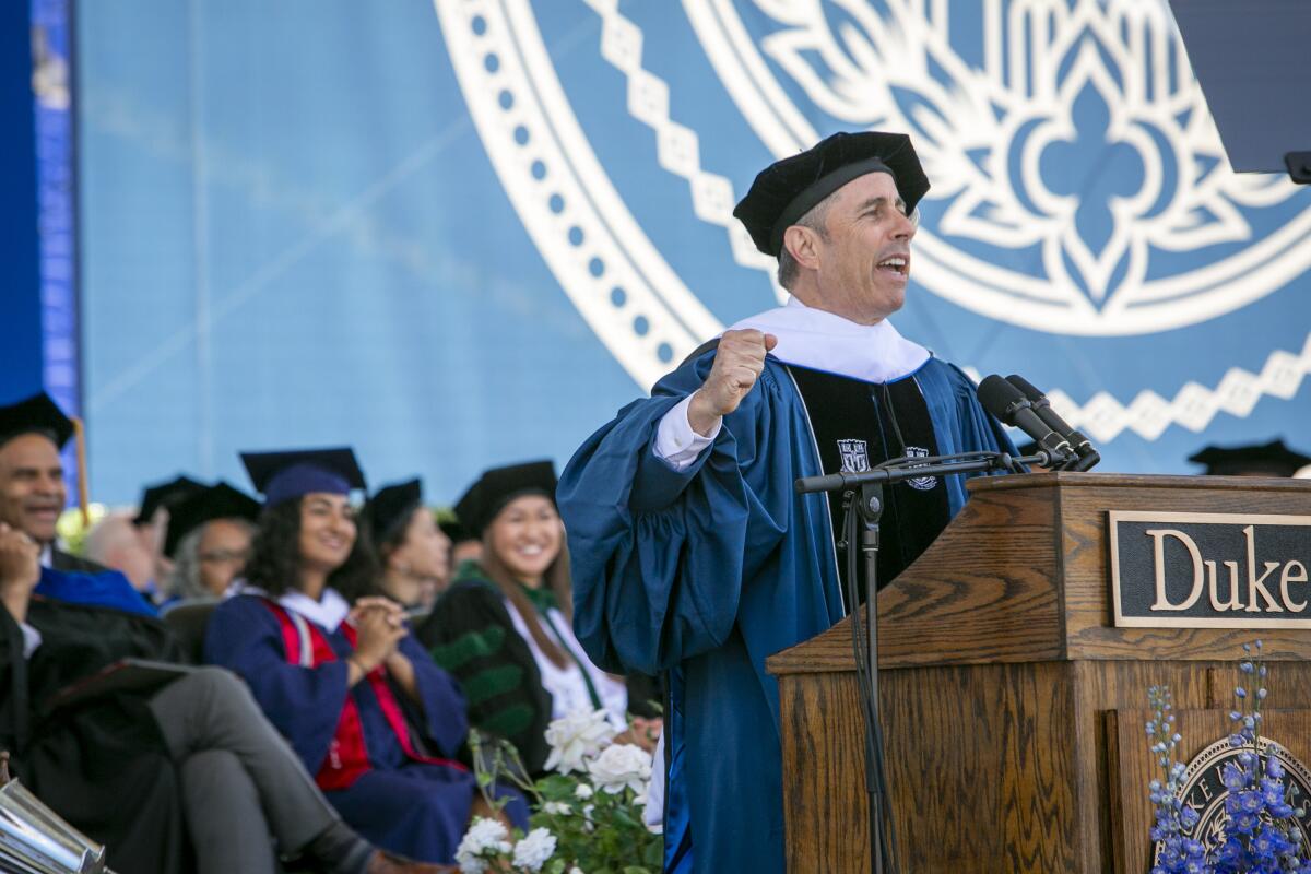 Jerry Seinfeld in a blue robe and graduation cap standing behind a wooden lectern that says "Duke"