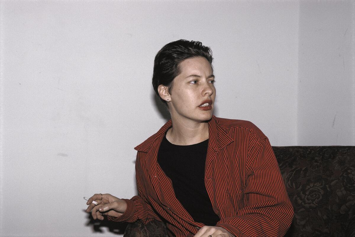 A photo of a woman with short hair, wearing a red and black striped collared shirt over a black tee, holding a cigarette.