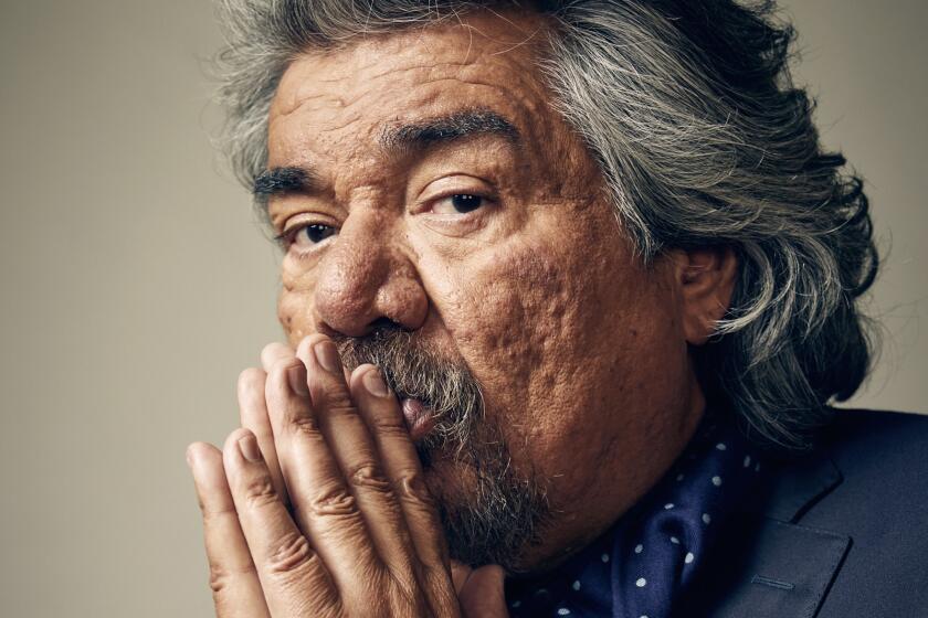 A vertical portrait of George Lopez holding his hands together in front of his mouth