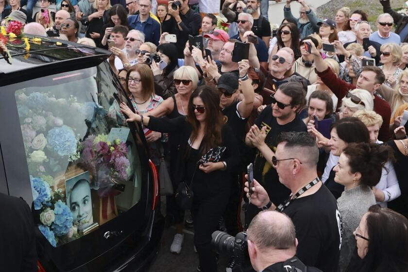 A crowd of people in black surround a hearse filled with pink and blue flowers and a picture of late singer Sinéad O'Connor