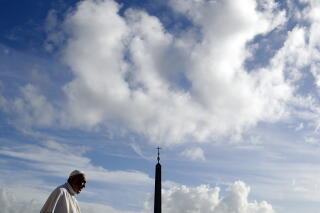 Pope Francis in the foreground while puffy white clouds fill the blue sky above and behind him and a cross sits atop a spire.
