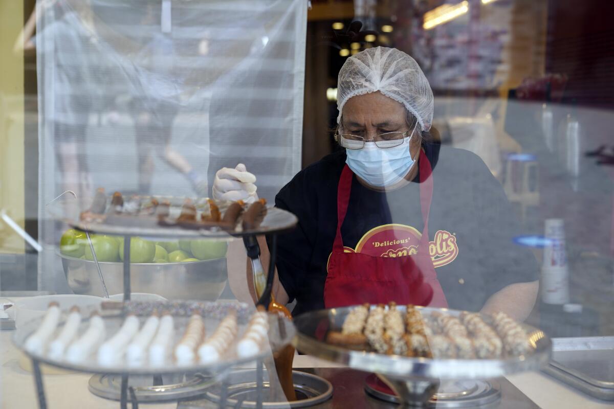 A masked and gloved worker in a red apron is seen behind a shop window with plates of desserts