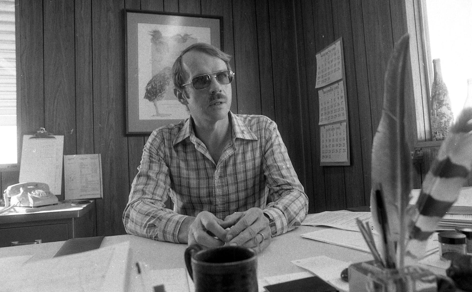 A scientist in a checkered shirt and sunglasses sits at a desk.