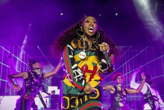 Missy Elliott in a yellow, green and red jumpsuit on stage holding a microphone. People in black outfits dance behind her