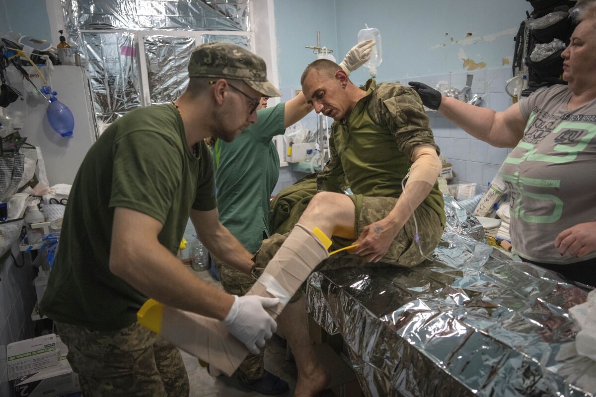 A man in a camouflage cap holds the bandaged leg of a man in fatigues seated on an examination table 