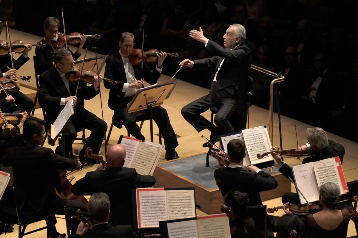 A man in tails sits on a stool and lifts a baton as he conducts an orchestra.