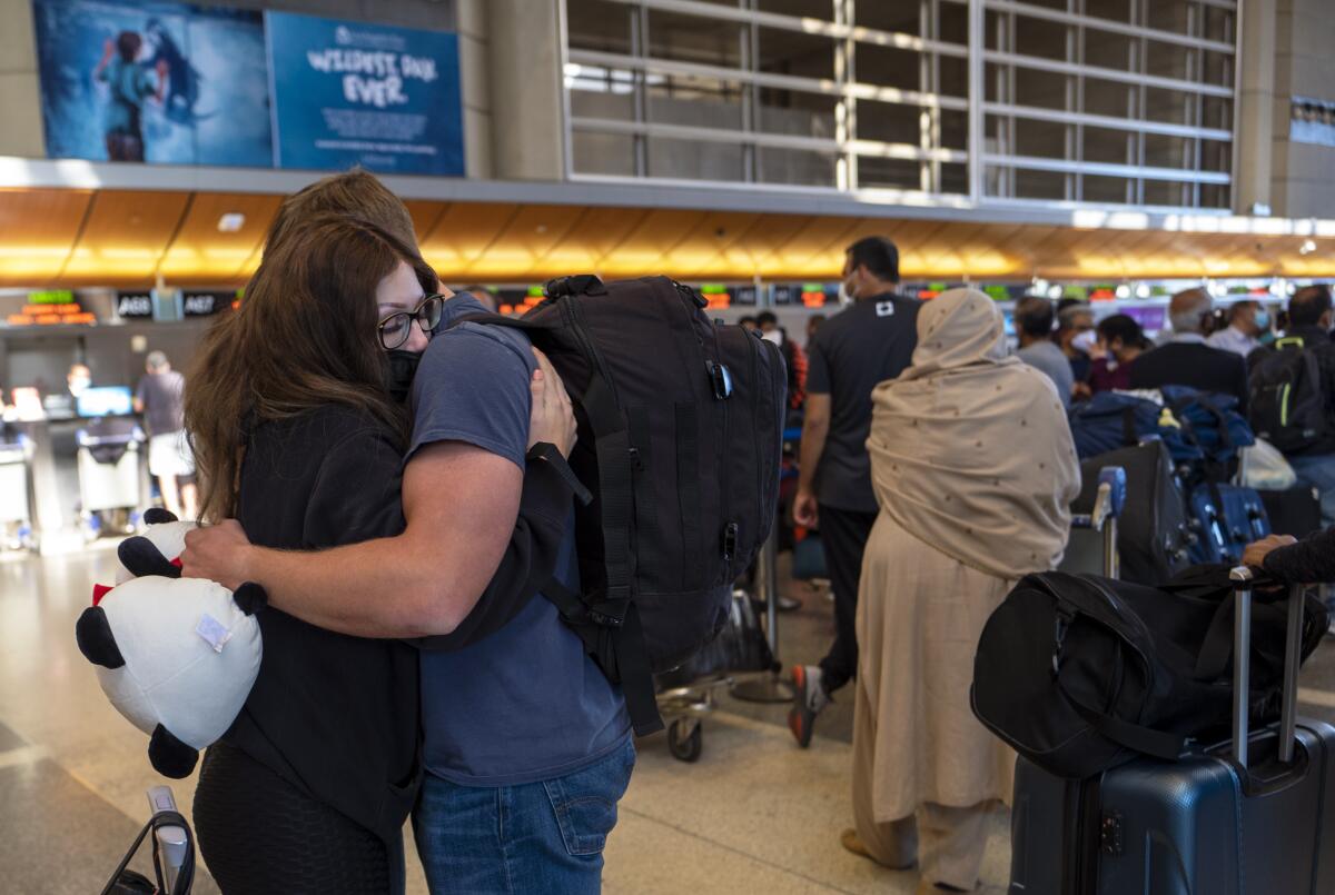A woman hugs a man wearing a backpack near lines of travelers in an airport terminal