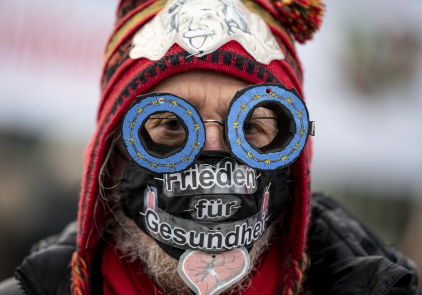 A participant of the nationwide day of action "Disarmament instead of rearmament" is standing in front of the Federal Chancellery wearing handmade glasses with painted barbed wire and a mouth and nose protector with the inscription "Peace for Health" in Berlin, Germany, Saturday, Dec. 5, 2020. Hundreds of peace protesters formed a human chain outside the German parliament urging disarmament and an end to weapons exports. About 300 people formed the chain Saturday stretching from the parliament building to Chancellor Angela Merkel’s nearby office, demonstrating under the motto “peace, not armaments." (Fabian Sommer/dpa via AP)