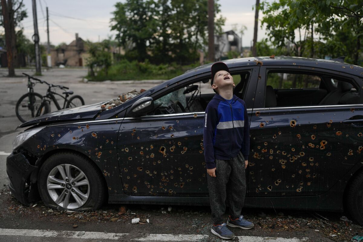 A child standing next to a damaged car looks up at a building destroyed.