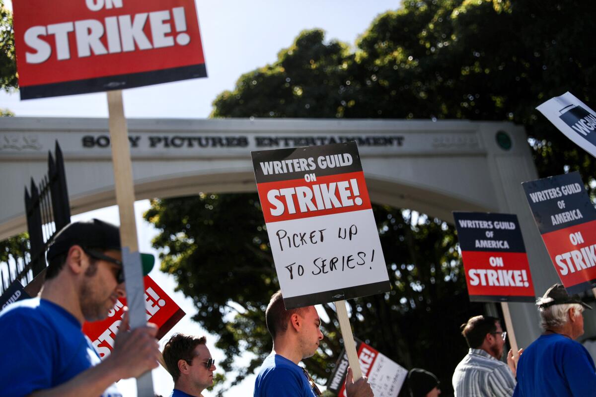 People hold up "Writers Guild on Strike!" pickets in front of an arched entrance outdoors.