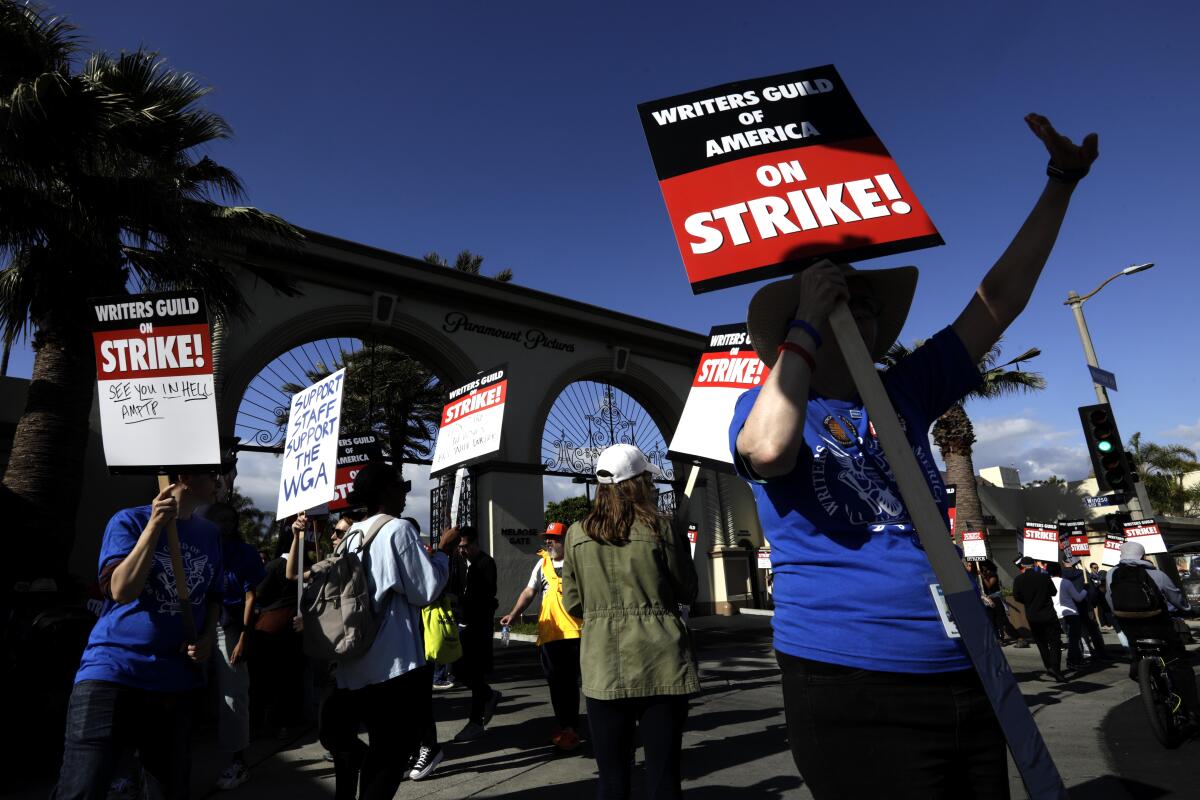 A group of people hold up signs about a writers strike outside a studio gate.