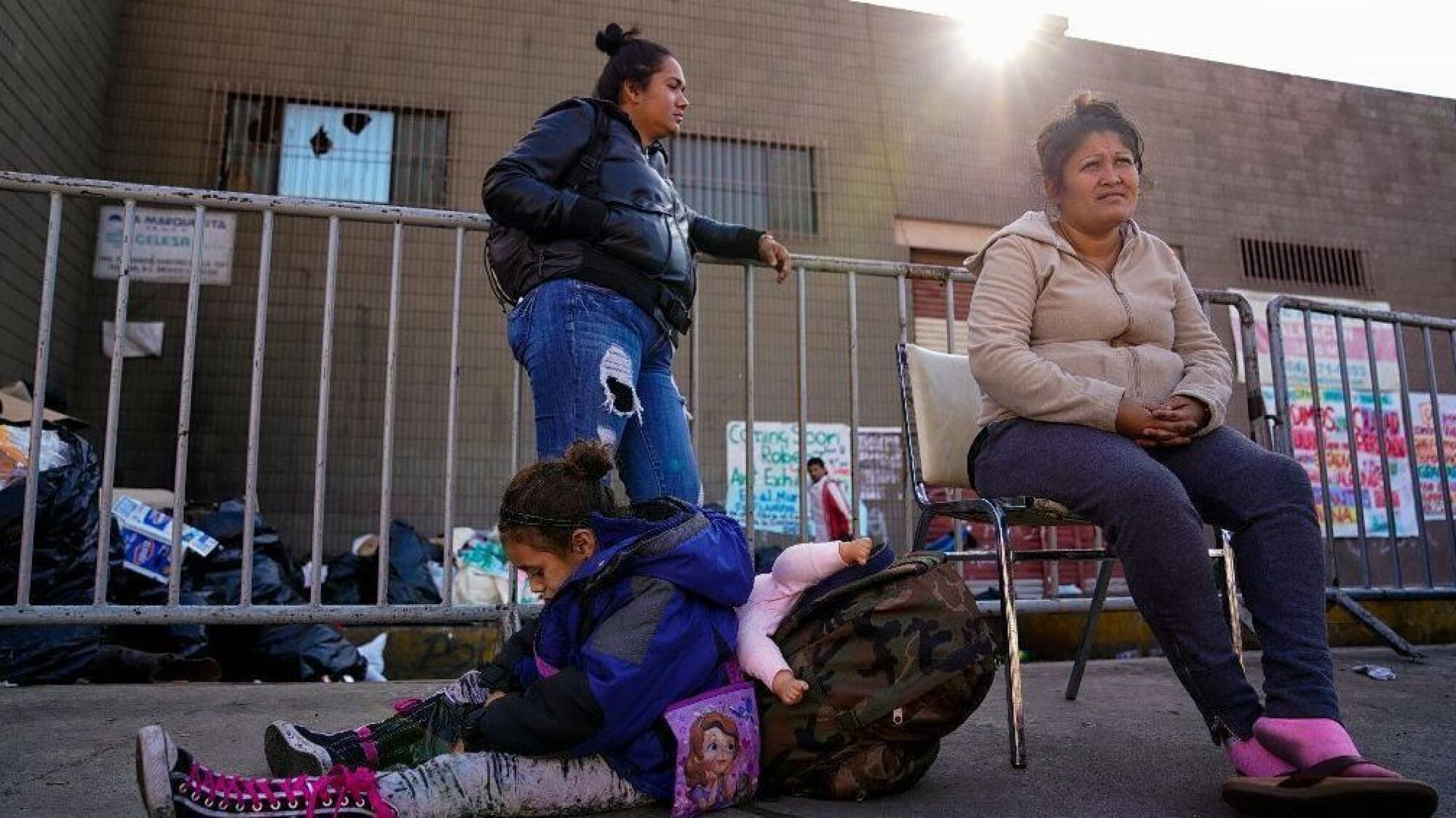 A woman and young girl sit while another woman stands, all three waiting by the port of entry to ask for asylum