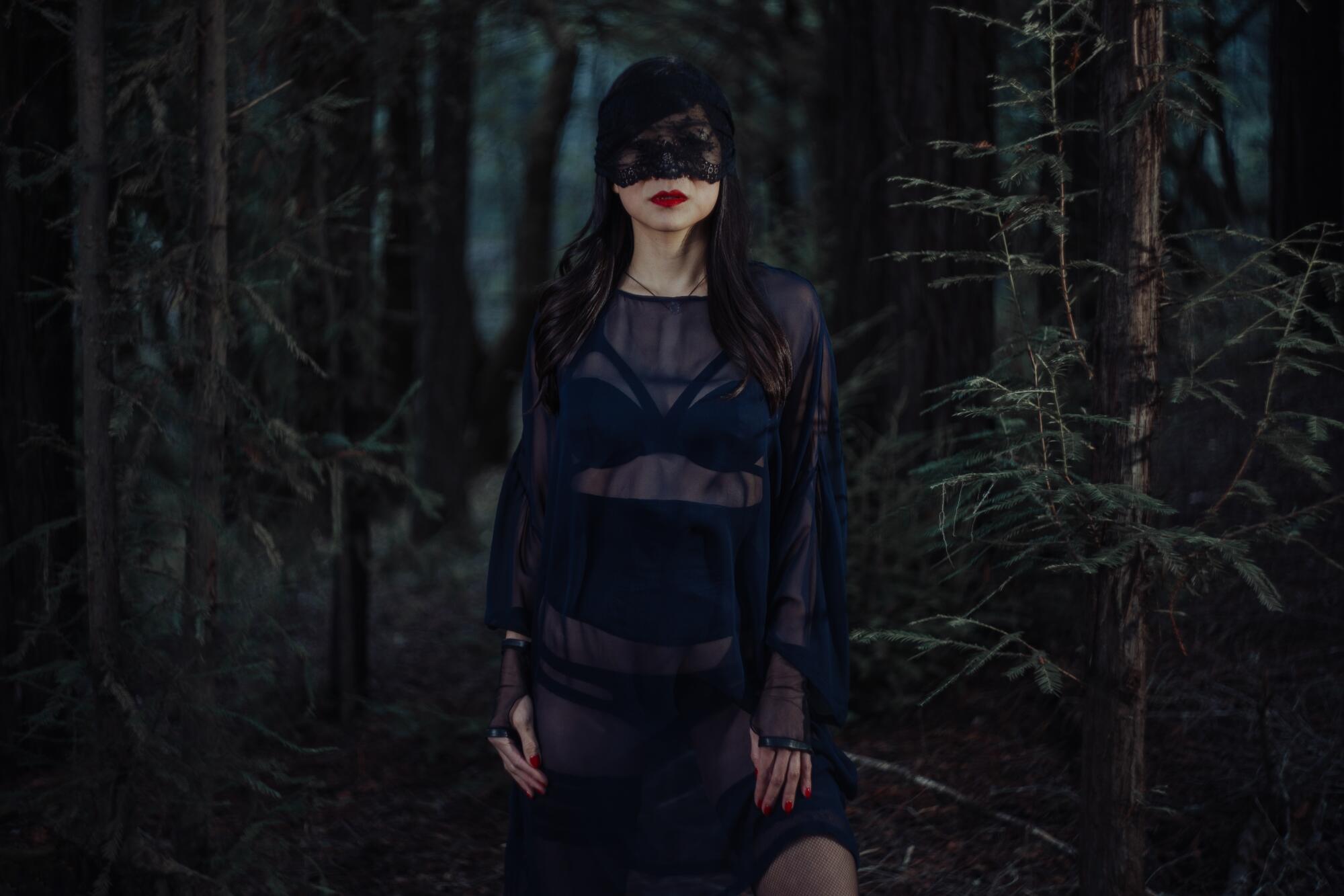 A woman in black lacy clothing and a black mask poses in a forest 