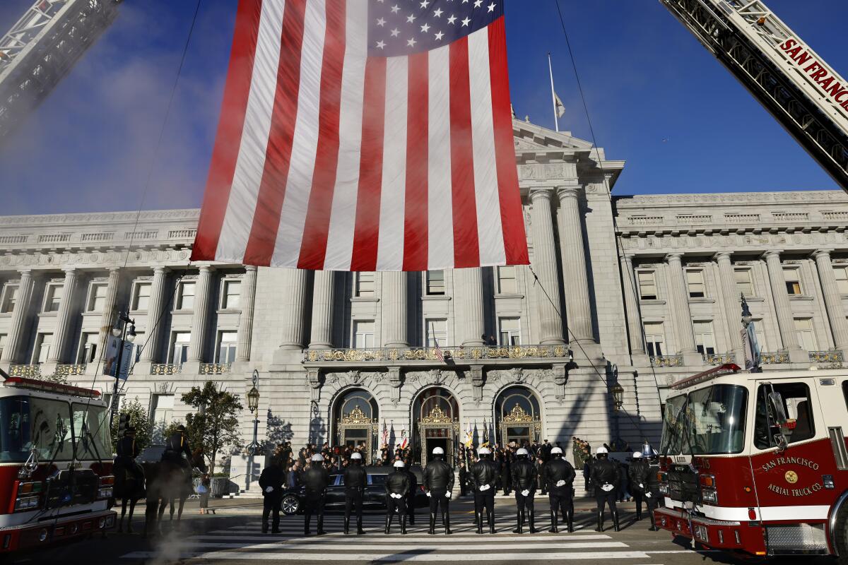 A large American flag held by cranes frames police and firetrucks lined up by a black hearse outside San Francisco City Hall