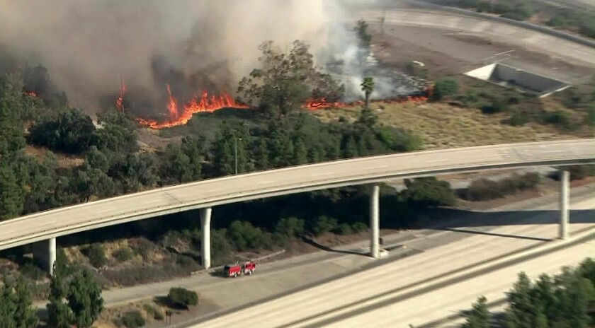 Behind elevated freeway lanes, a hillside is on fire; a firetruck is in the foreground. 