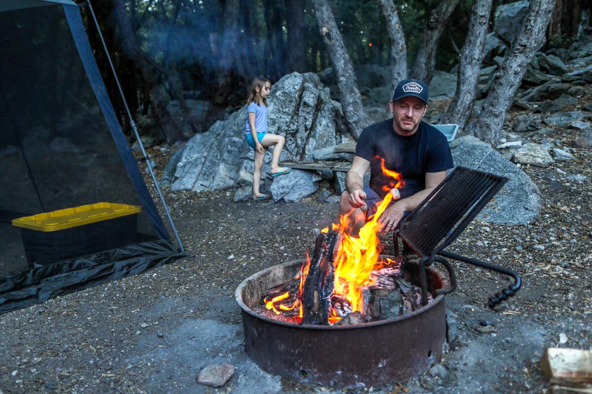 A man roasts marshmallows at a campfire while his daughter plays in the background by a tent.