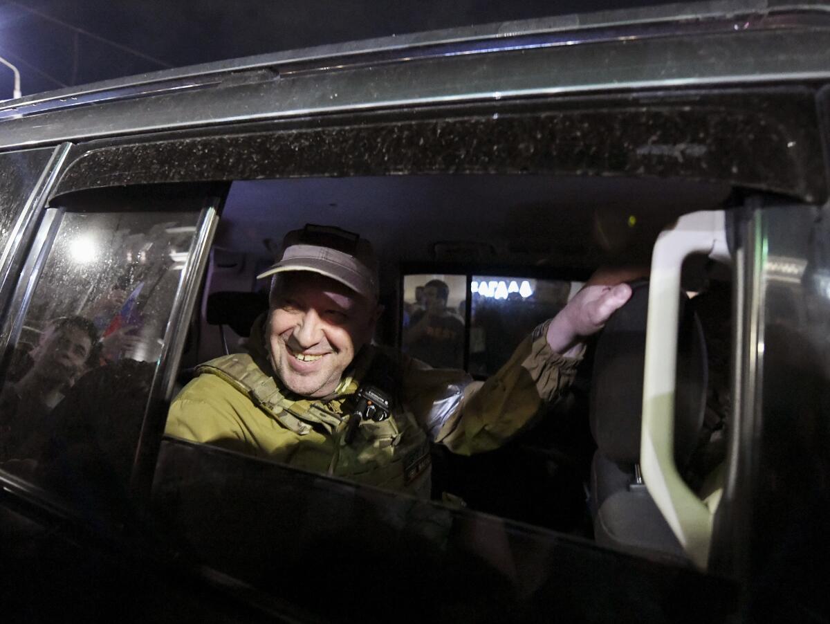 A man, wearing military jacket, smiles while seated inside a car, framed by the back-seat window 