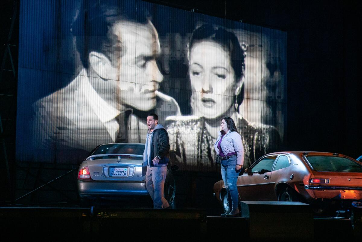 Two performers in casual contemporary clothes sing on a stage set that resembles a drive-in, complete with cars.