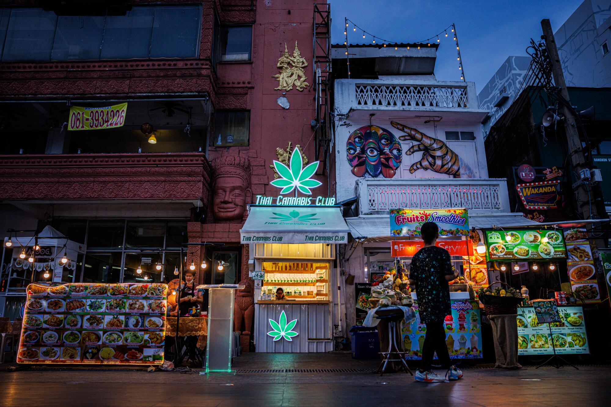 A person is seen in front of lighted storefronts, including one bearing signs shaped like marijuana leaves 