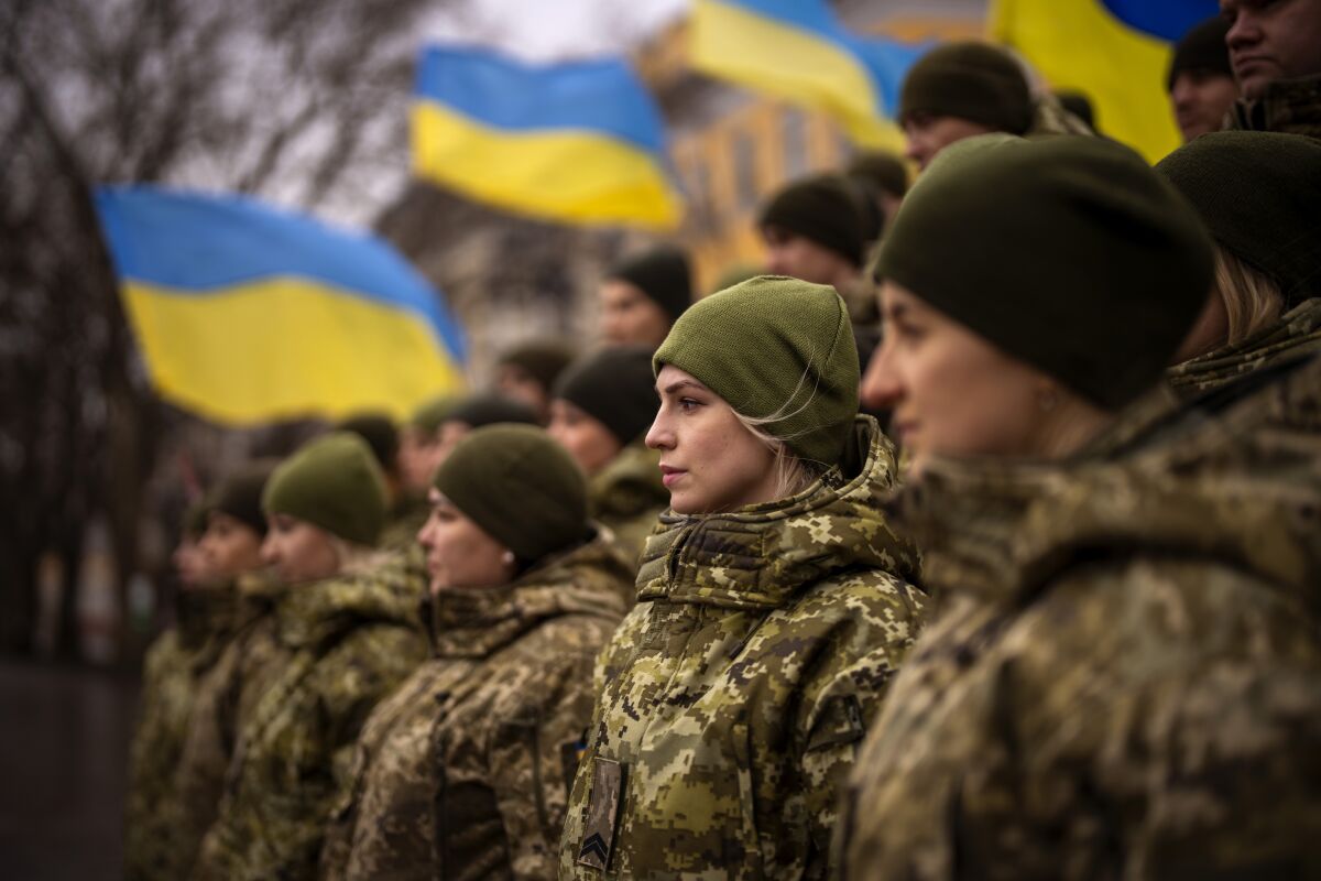 A group of female soldiers in camouflage stand near others and blue and yellow flags