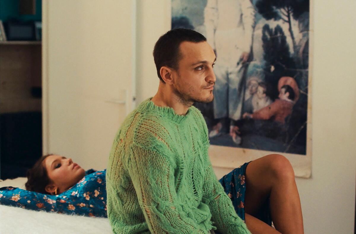 A man in a green sweater sits on the edge of a bed. A woman lies next to him.
