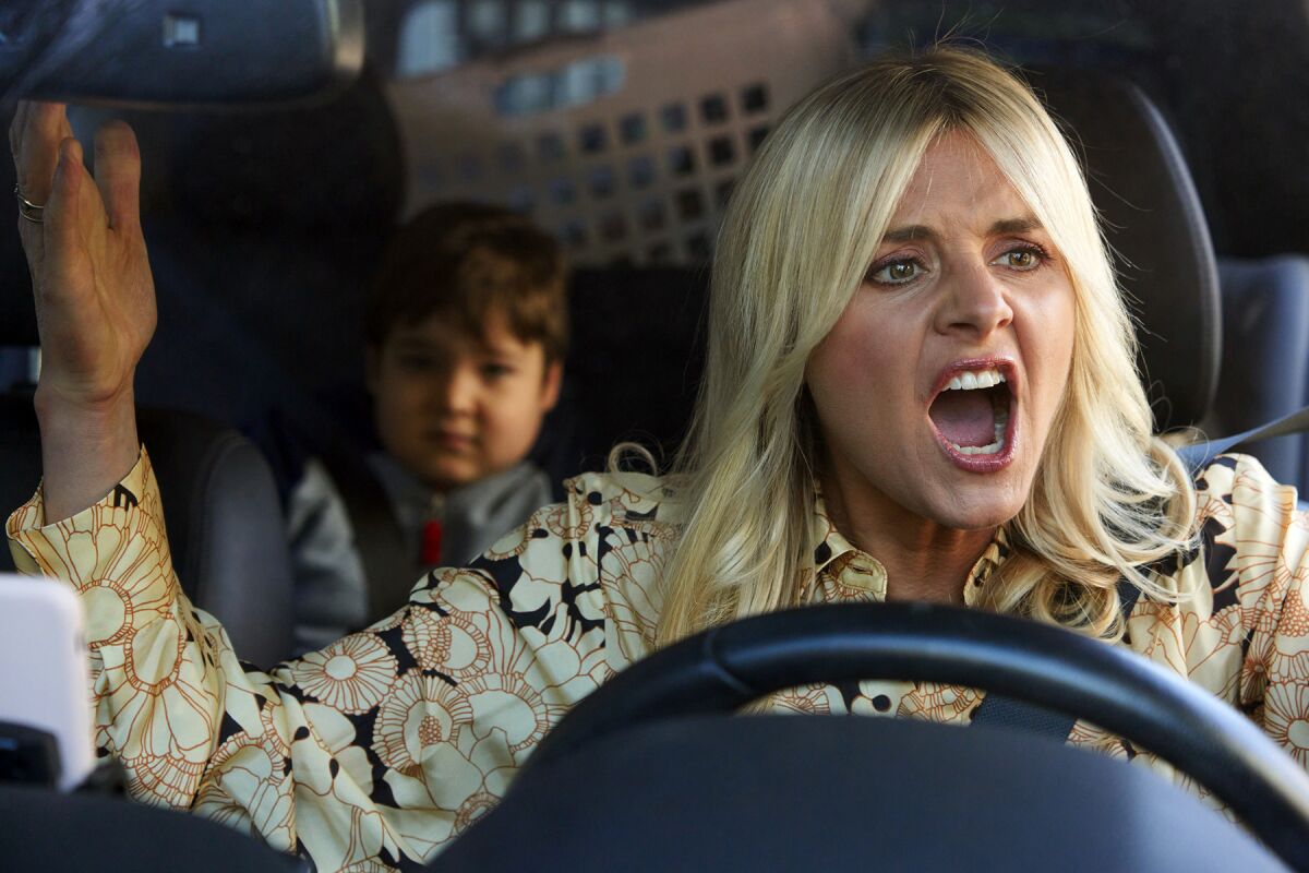 A blond woman behind the wheel of a car, with a child in the back seat, throws up her arms in anger.