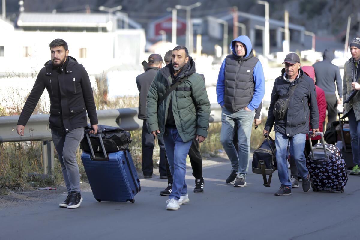Men in winter coats, some with luggage, walk on a sidewalk