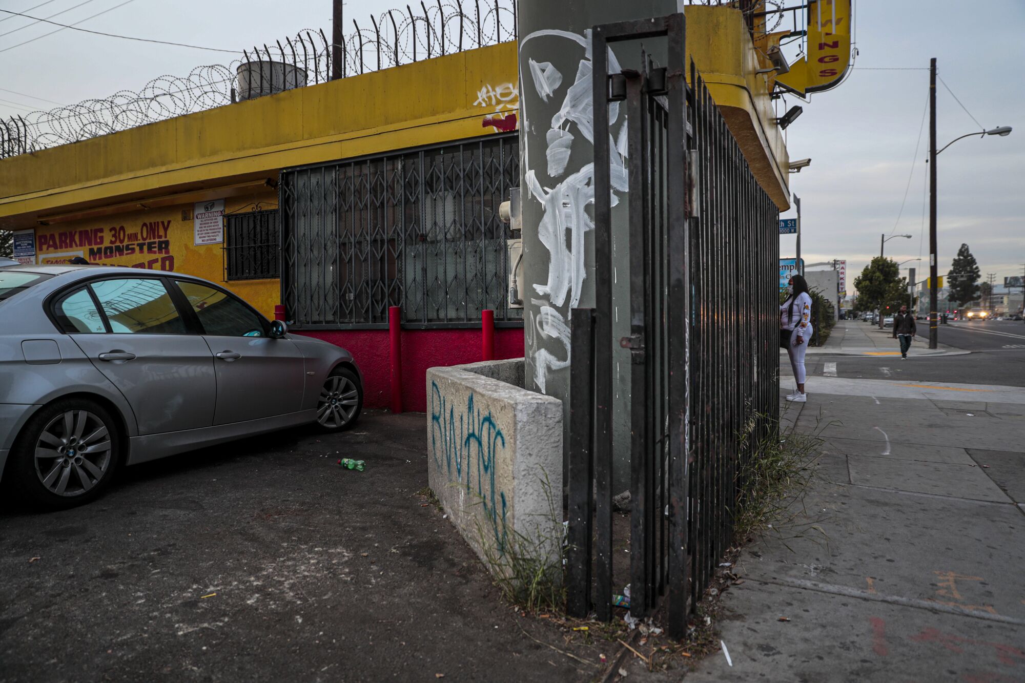 A burger joint sits in a parking lot with graffiti-tagged concrete barriers, bars on the windows and a security gate.