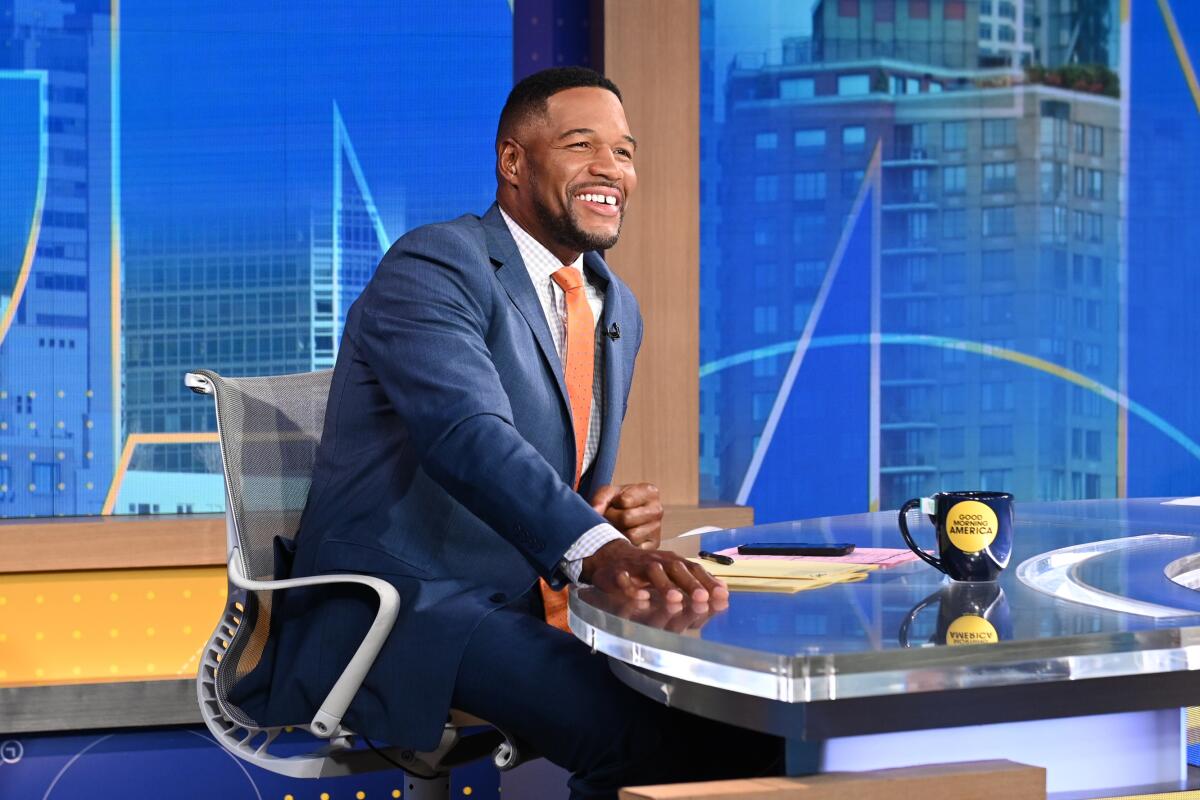 Michael Strahan sits at an anchor desk and smiles while wearing a blue suit and orange tie