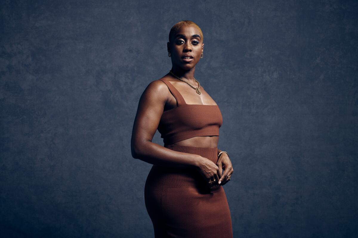 Lashana Lynch dressed in a brown skirt and matching midriff-baring top poses for a portrait.