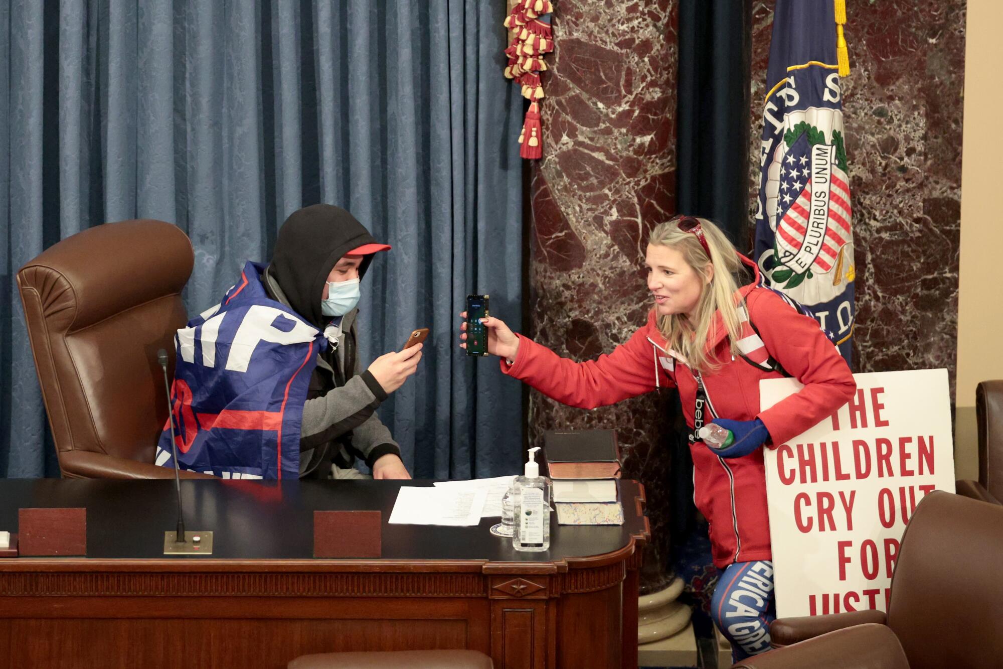 A man wearing a Trump flag sits in the Senate chamber. He and a woman look at their phones.