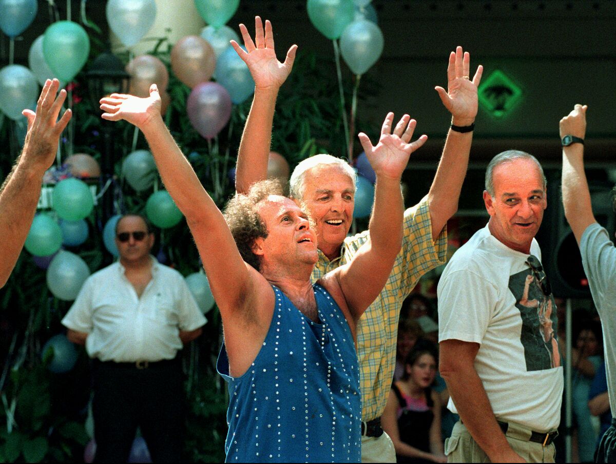 A man in a tank top with his arms raised leads a group of older people exercising