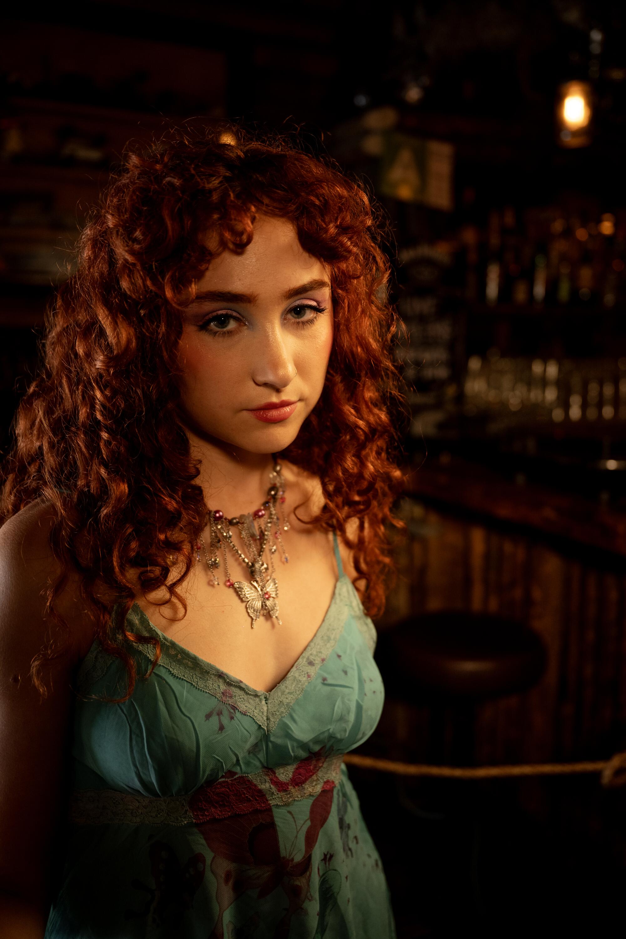 A red-headed woman in a green top and necklace looks downward pensively.