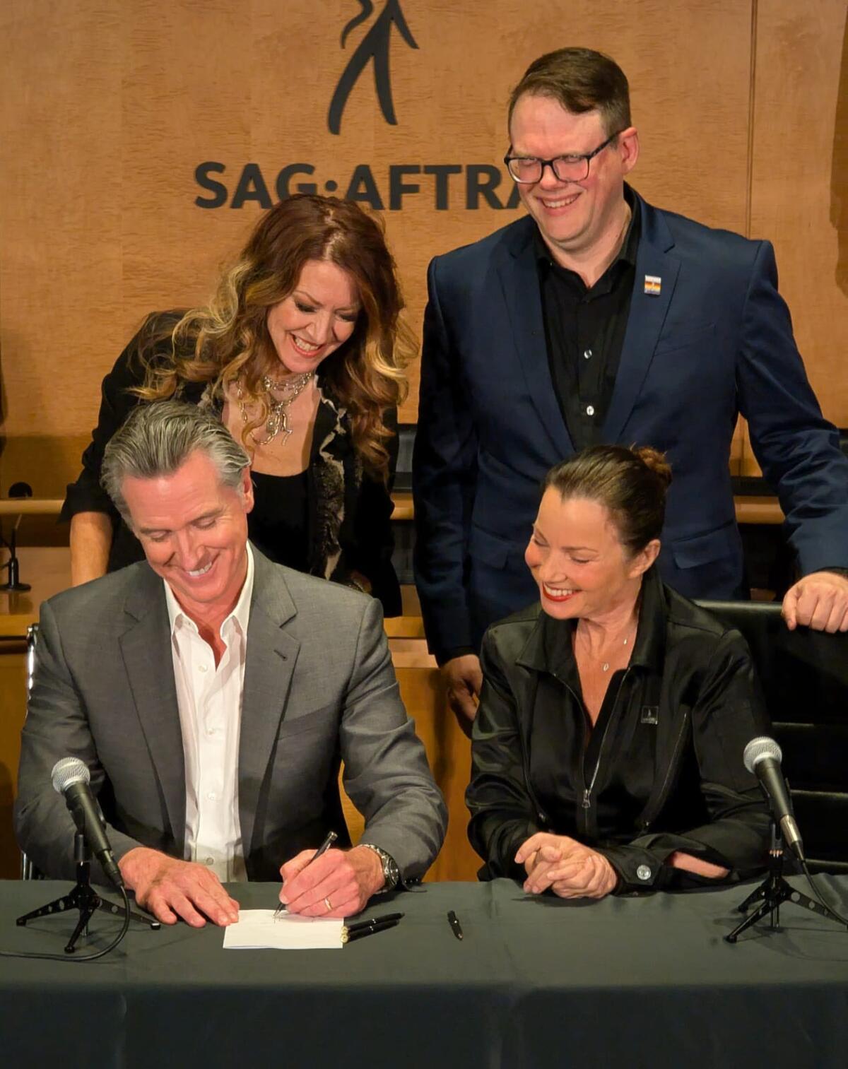 A man in a gray suit smiles as he signs documents, while two women and a man look on