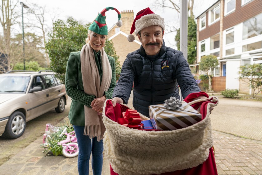 A woman in an elf hat and a man in a Santa hat hold out a bag of toys