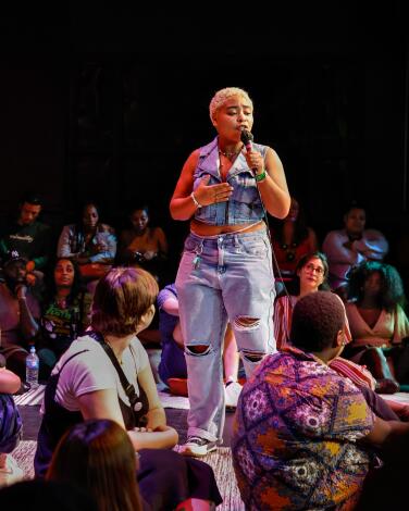 A woman performs at an open mic poetry slam at Da Poetry Lounge, surrounded by people sitting on the floor.