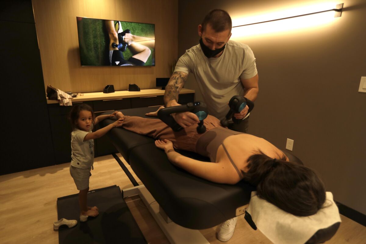 A man gives a woman a massage using Theragun massage devices. A young girl stands next to the massage table.