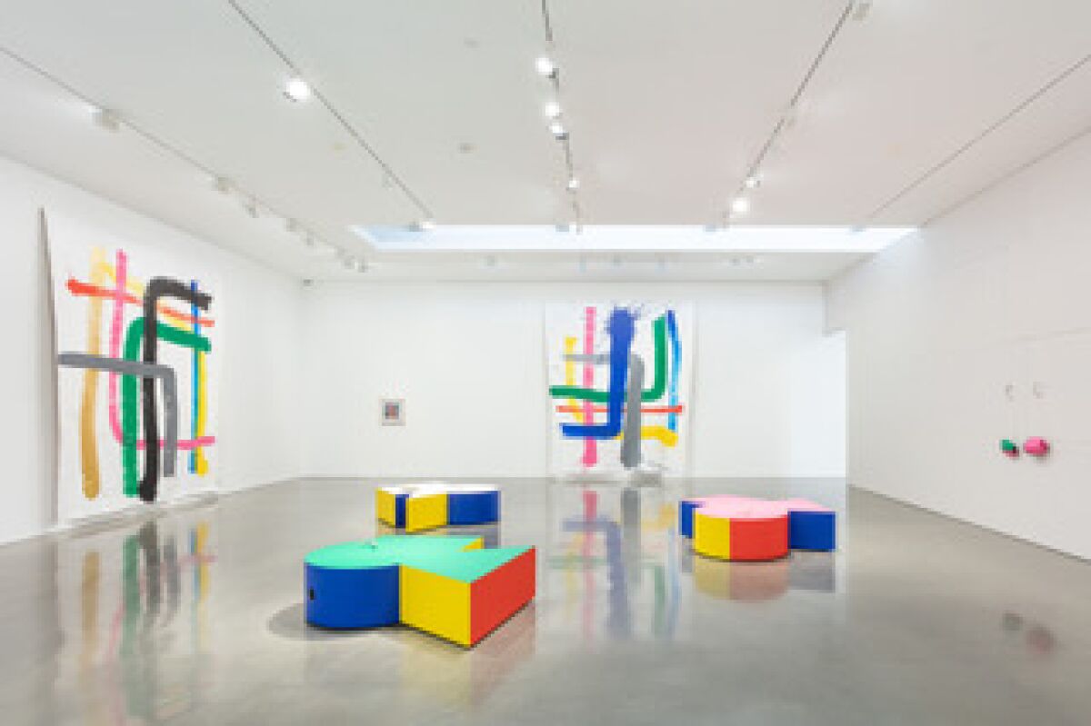 Colorful paintings on lean against the wall, with colorful boxy sculptures in the middle of the floor 