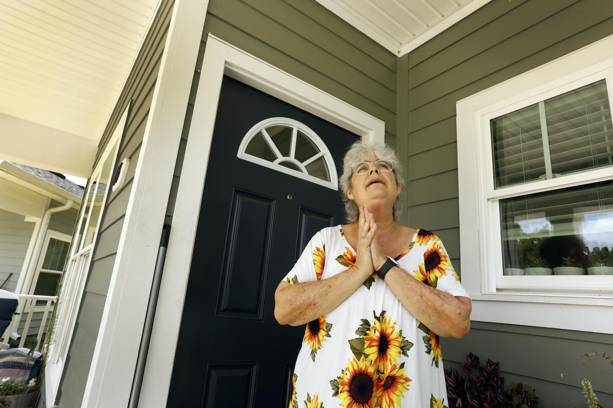 A woman with gray hair, wearing a flowery dress, holds her palms together as she stands outside the dark blue door of a home